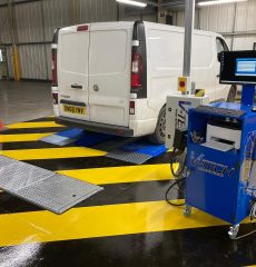 White van on a wheel alignment bay with vehicle testing equipment in a garage workshop.